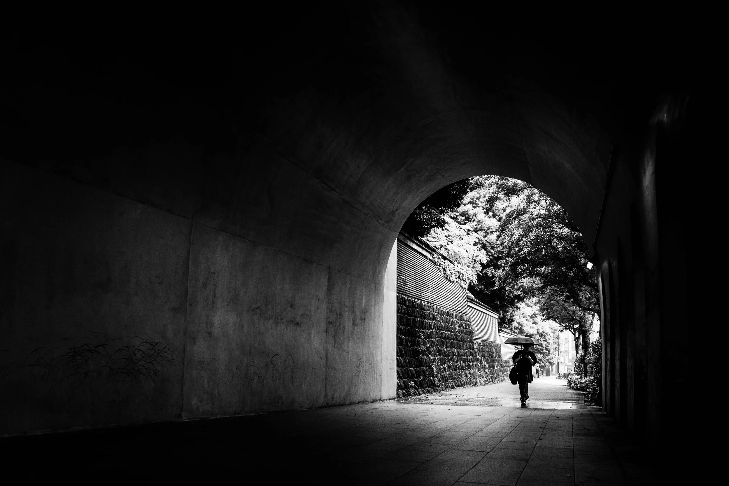 Schwarz-weiß-Foto einer Person mit Regenschirm, die durch einen Tunnel nach draußen geht, mit Bäumen im Hintergrund.