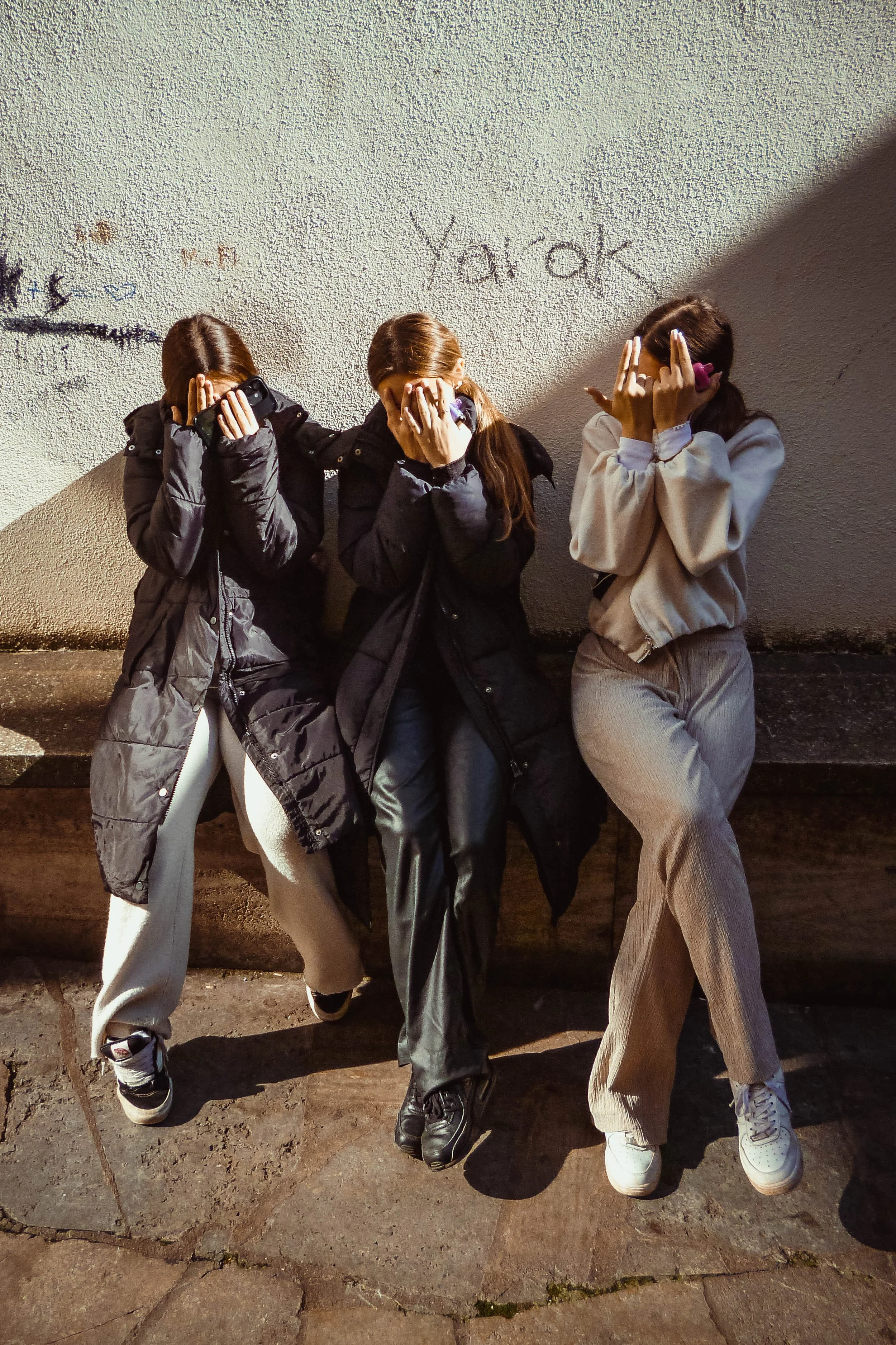 Drei Mädchen sitzen auf einer Bank, haben die Hände vor das Gesicht und tragen Winterjacken. Sie lehnen an einer wand mit Graffiti.