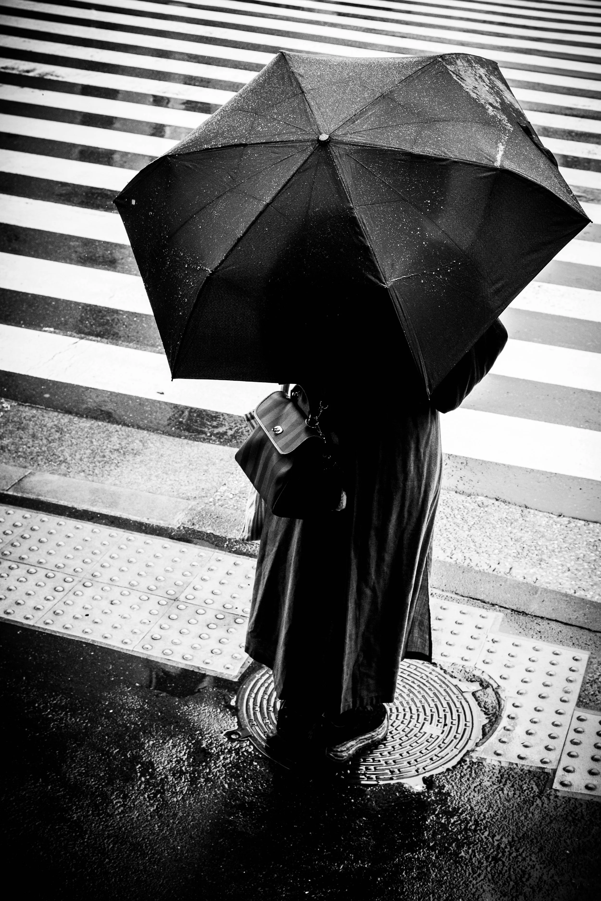Eine Person mit langem Mantel hält einen schwarzen Regenschirm auf einer nassen, dunklen Straße und wartet an einer Kreuzung.