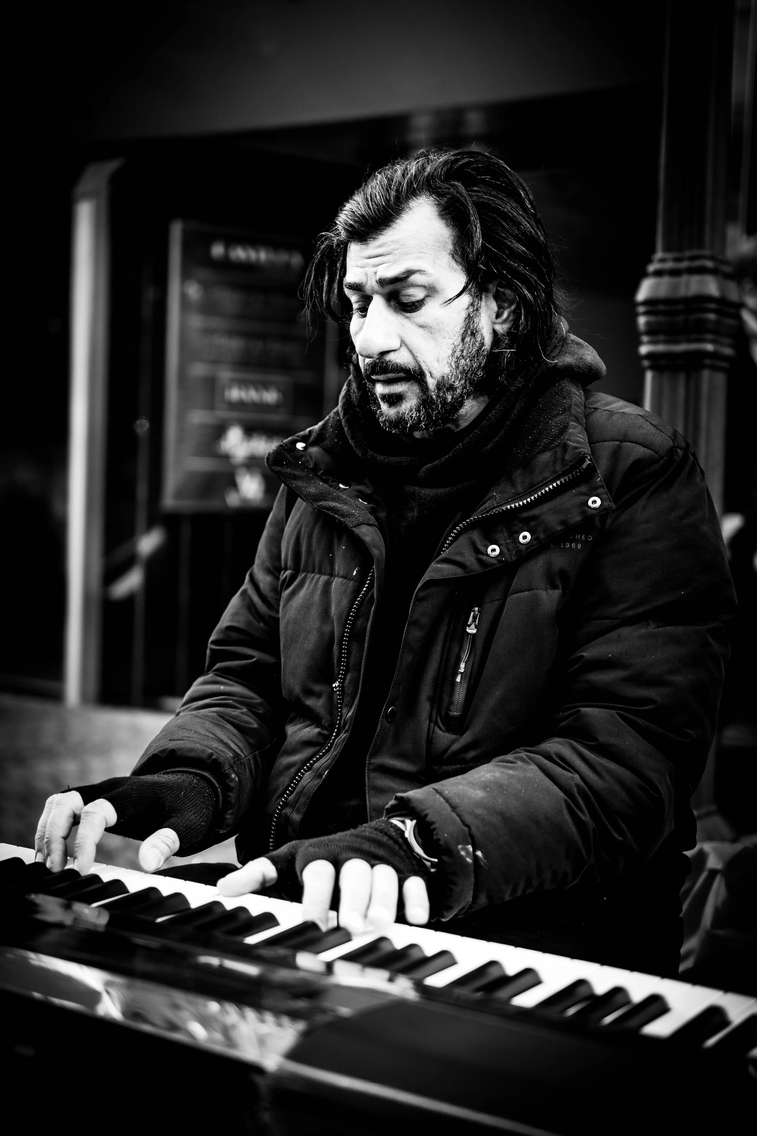 Ein Mann spielt Klavier in schwarz-weiß, trägt eine dunkle Jacke und Handschuhe, hat dunkle, schulterlange Haare und einen Bart.