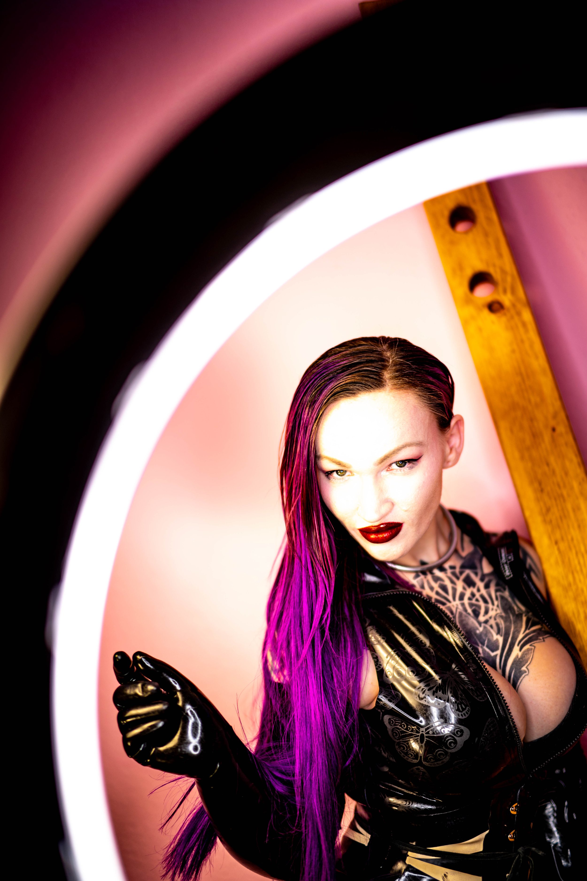 Frau mit langem, gefärbtem lila Haar und Tätowierungen, trägt schwarzes, glänzendes Outfit und Handschuhe, posiert vor einem runden Lichtrahmen in einem Raum mit pinker Beleuchtung.