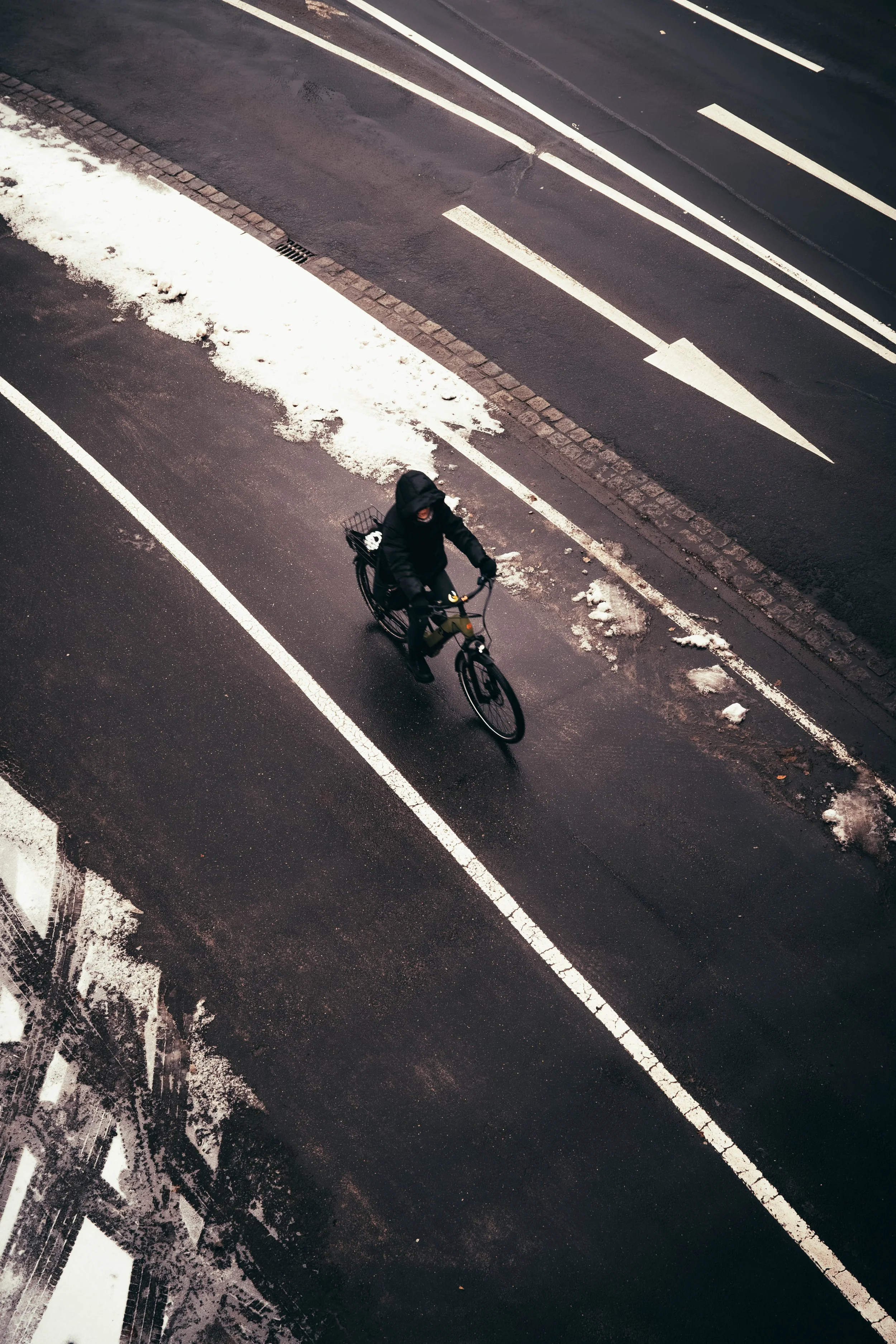 Ein Mensch fährt mit dem Fahrrad auf einer nassen, leeren Straße im Winter, mit Schneeresten am Straßenrand.