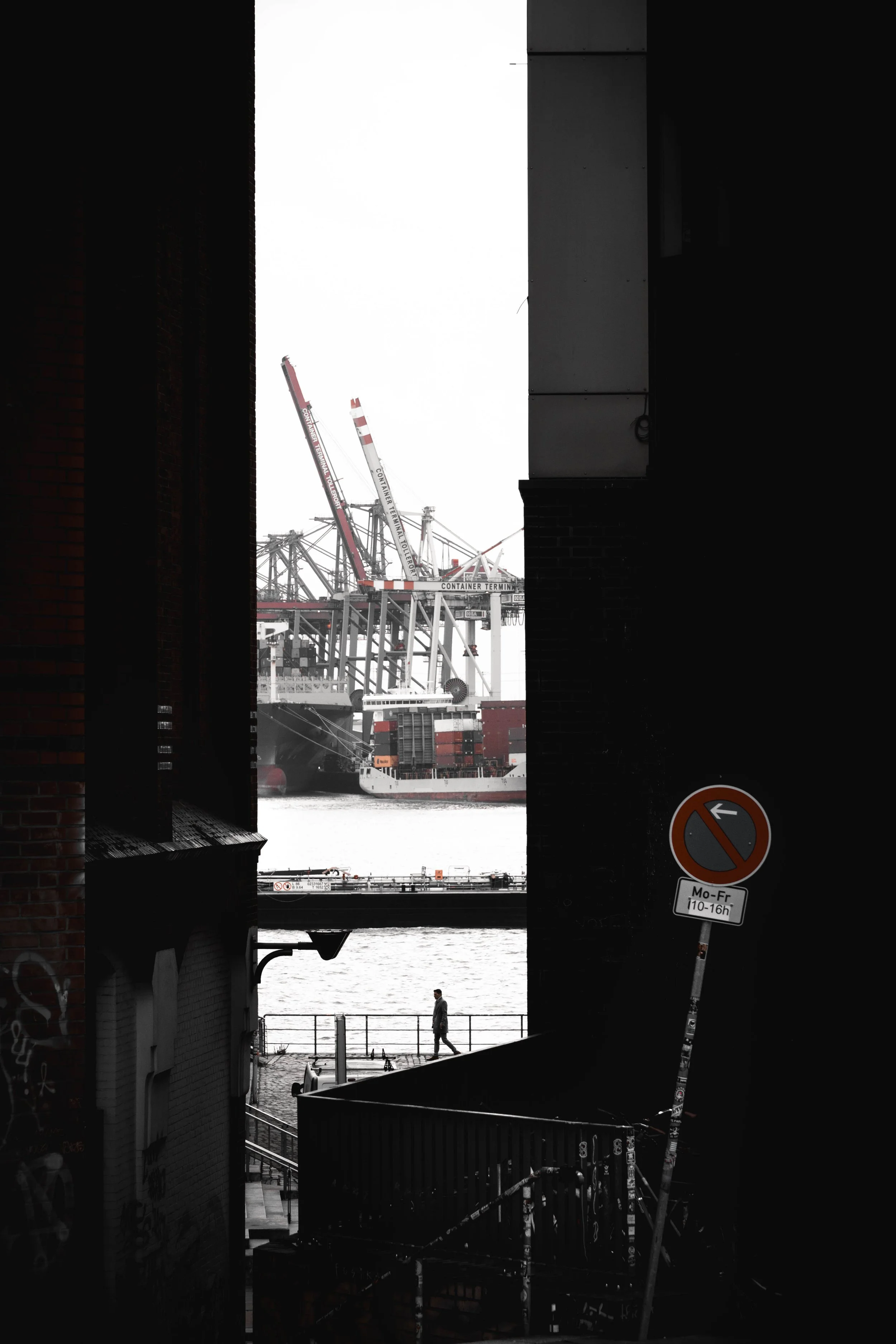 Blick durch eine enge Gasse auf einen Hafen mit Containerkränen im Hintergrund, eine Person läuft am Wasser entlang, ein Verkehrsverbotsschild ist sichtbar.