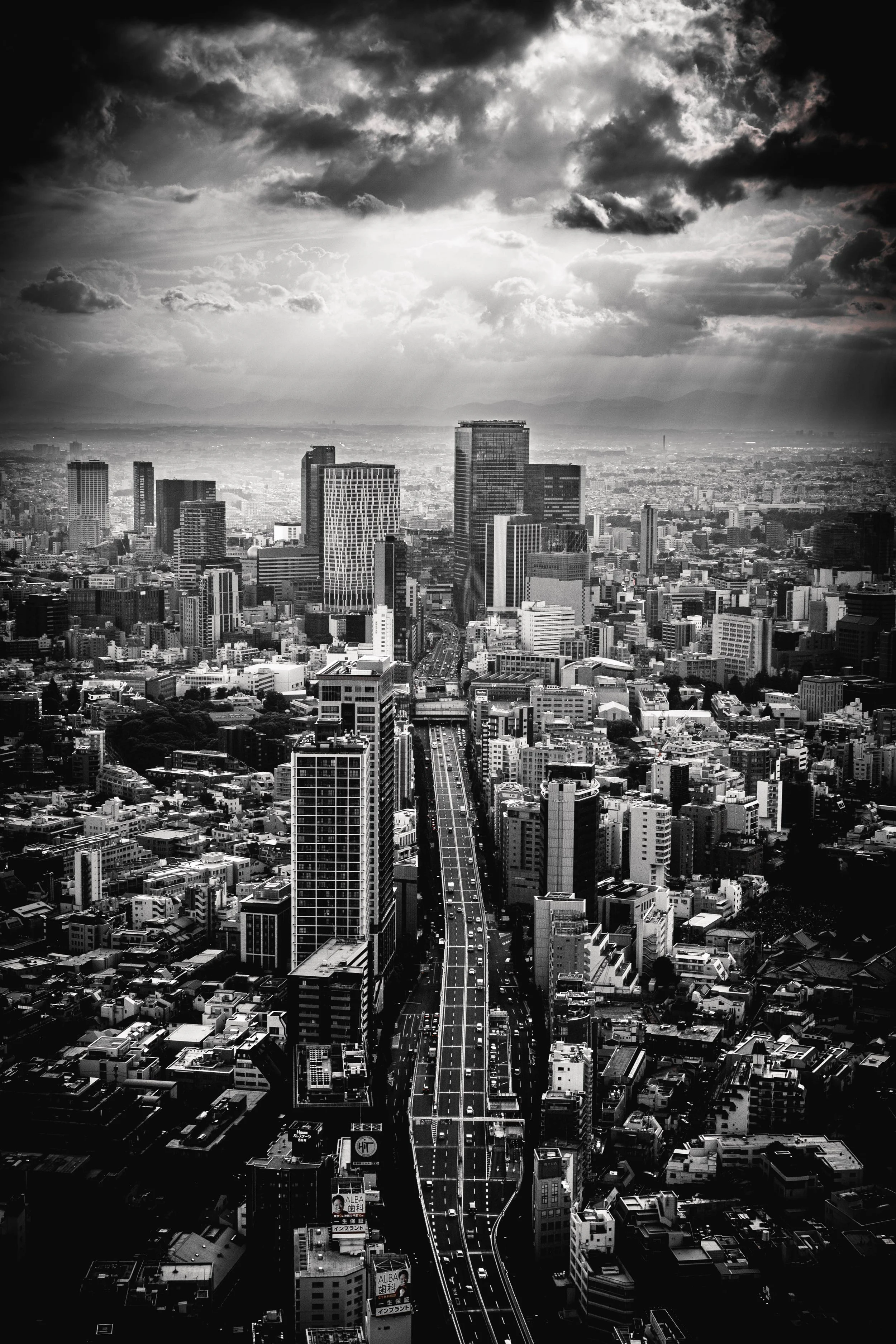 Schwarz-Weiß-Foto einer japanischen Stadt mit Hochhäusern, einem Straßenmuster im Vordergrund und einer bewölkten Himmelsszene im Hintergrund.