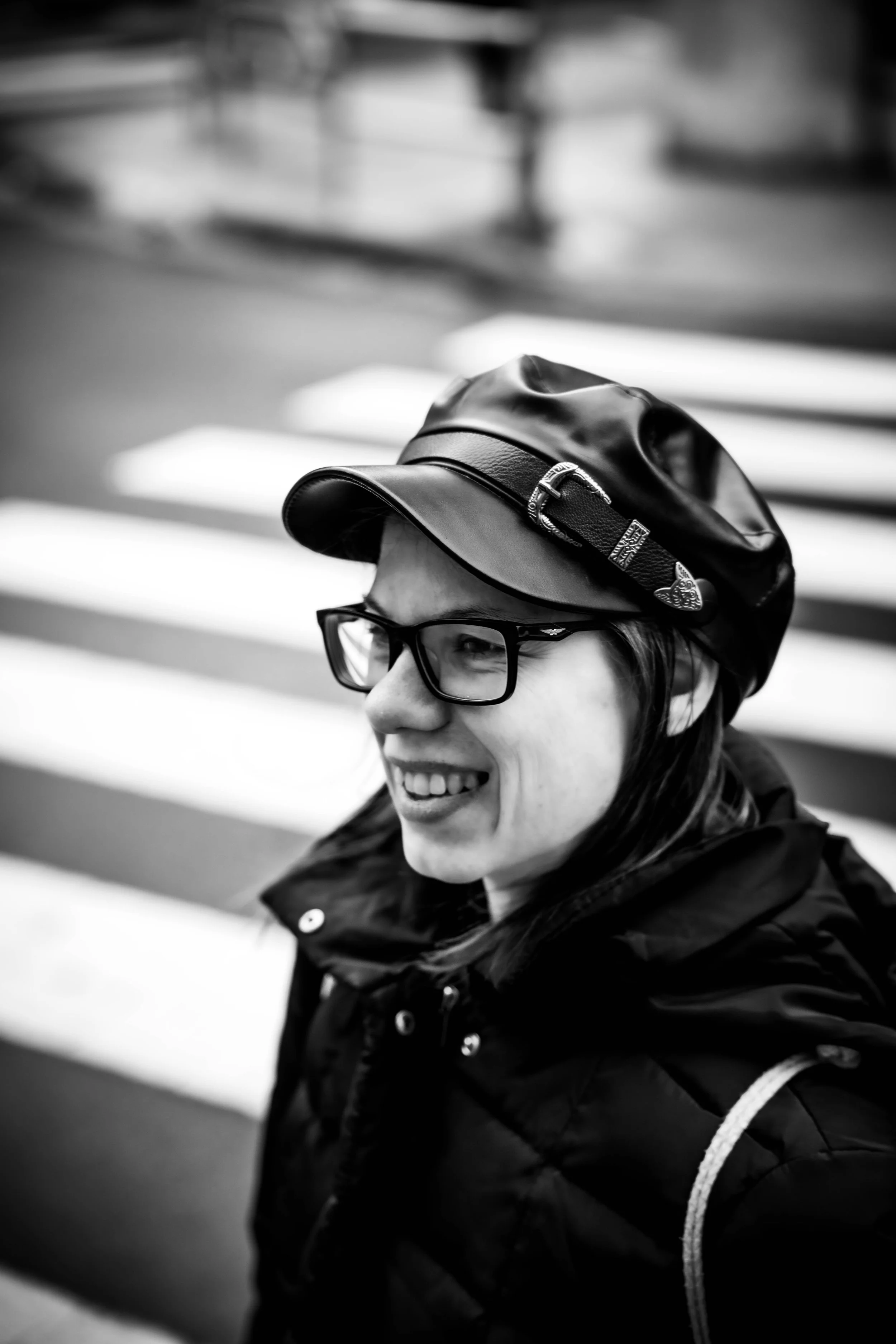 Nahaufnahme einer lachenden Frau mit Brille, trägt eine Lederkappe und eine wetterfeste Jacke, im Hintergrund einer Straßenkreuzung.