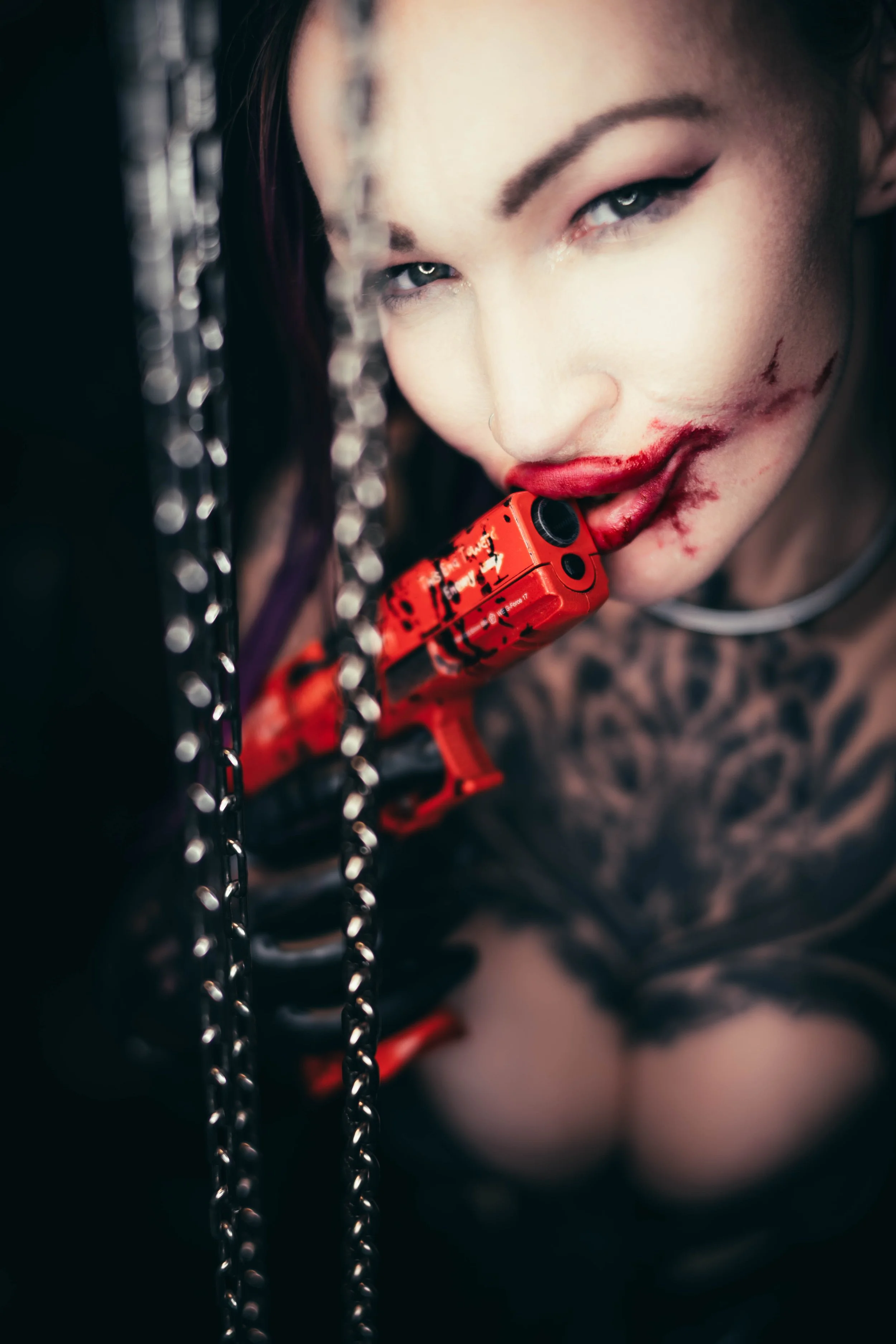Frau mit Make-up und blutigem Gesicht hält eine rote Pistole in den Zähnen, umgeben von Ketten.