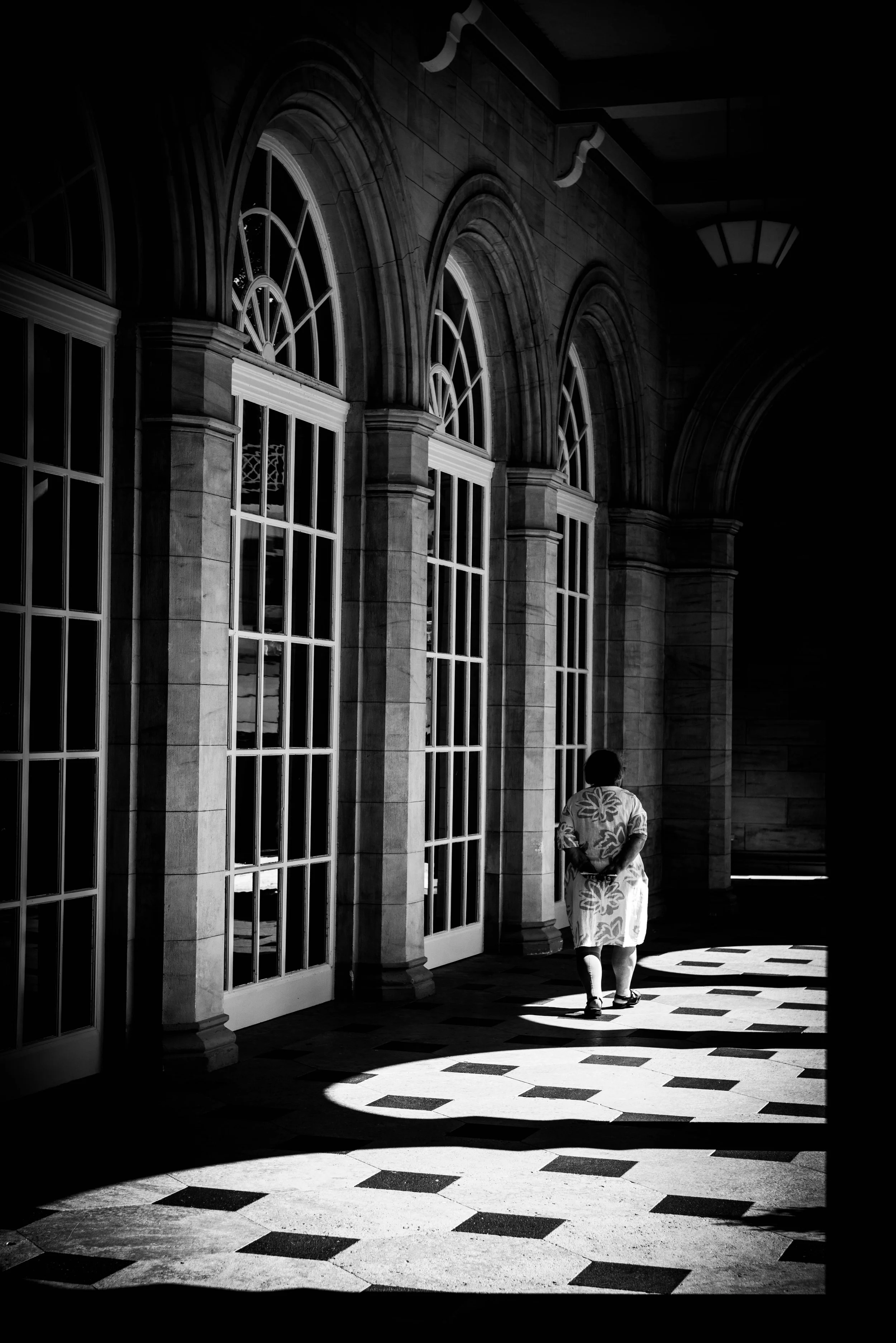 Eine Person in einem passenden Kleid geht durch einen Flur mit großen, bogenförmigen Fenstern und einem Schattenmuster auf dem Boden.