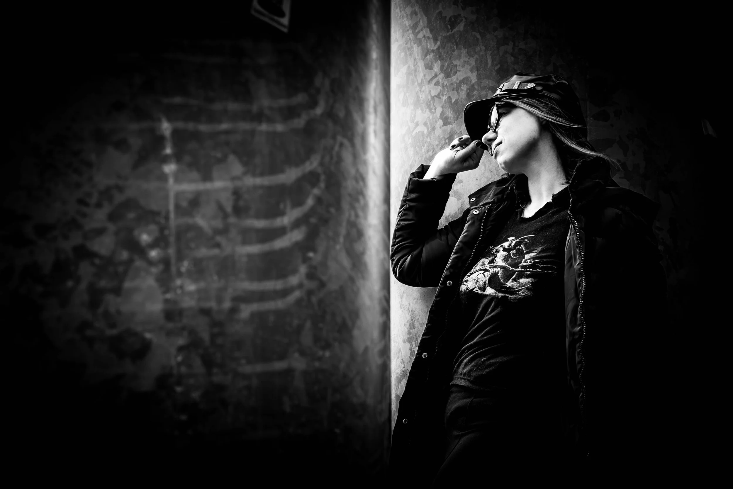 Schwarz-weiß-Foto einer lächelnden Frau mit Brille, Hut, dunklem Jacke und T-Shirt, die an einer Wand lehnt.