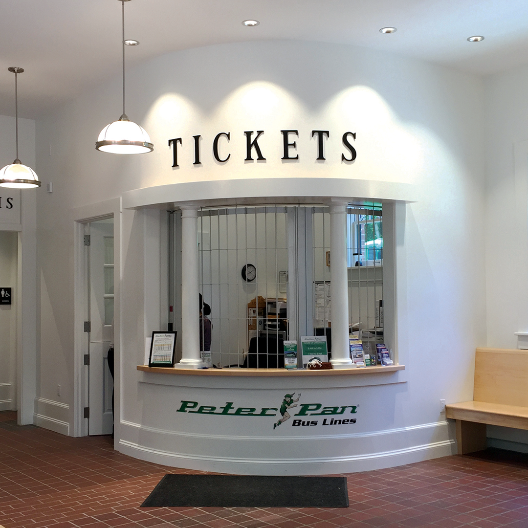 Bus ticket counter with a sign reading 'TICKETS' on a white wall, featuring the Peter Pan Bus Lines logo. The counter has two white columns supporting a curved top, with brochures and a computer on the counter. There are hanging lights above and a bench to the right.