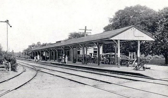 Black and white photo of a vintage train station with a few people and vehicles, including early 20th-century automobiles, on the platform and nearby tracks.