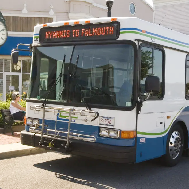 Public transit bus displaying route from Hyannis to Plymouth, parked on a city street with a woman sitting on a bench nearby.