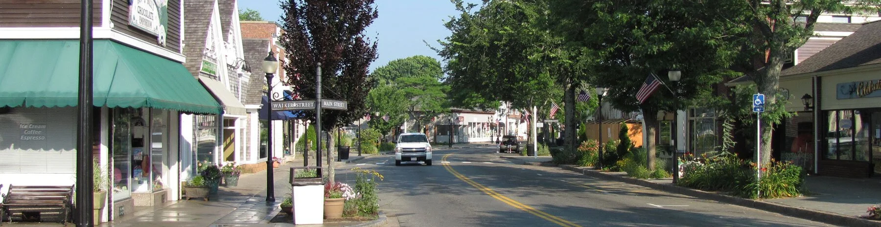 Main street in a small town with shops, trees, American flags, and a white vehicle driving down the street.