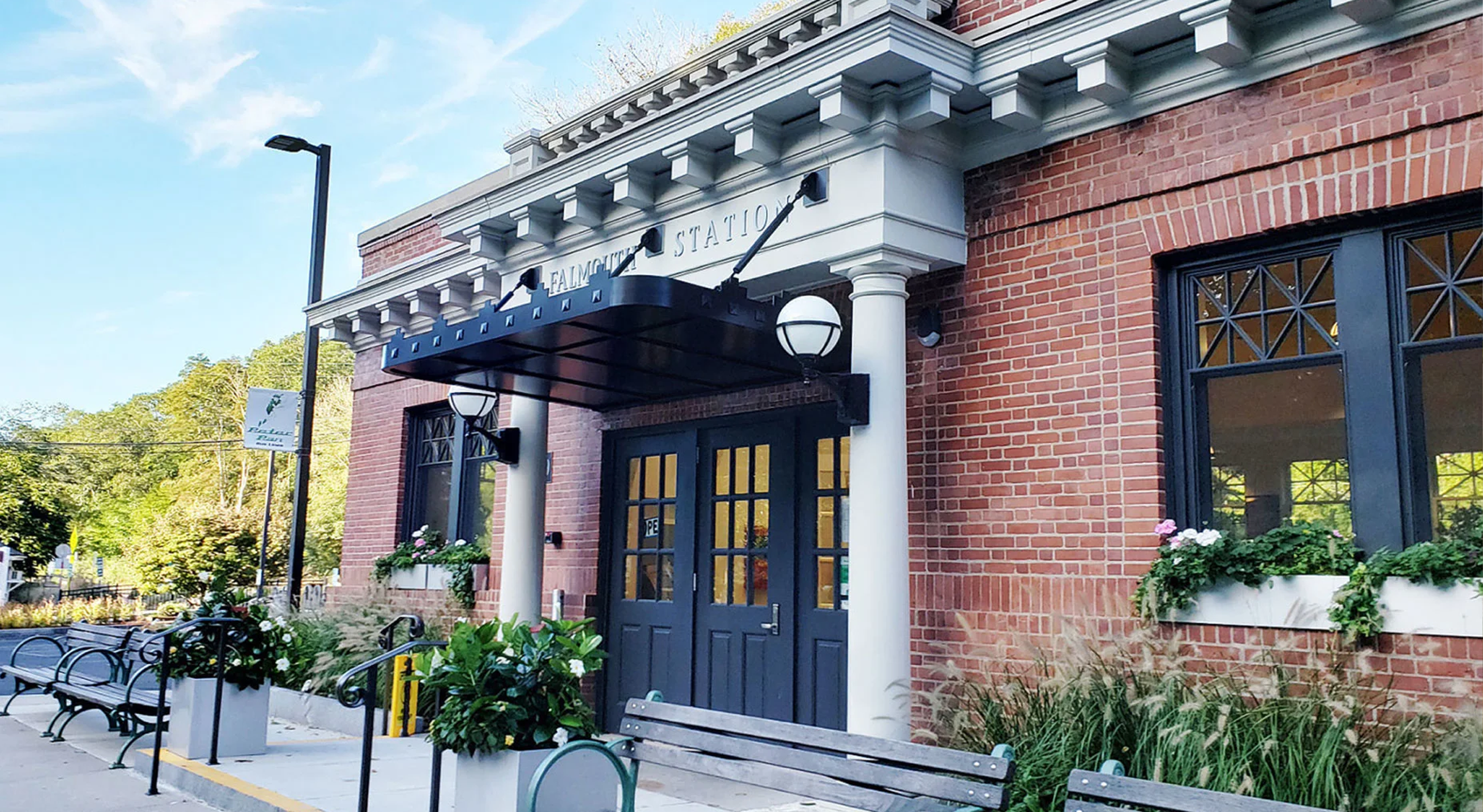 Exterior of a brick building labeled 'Falmouth Station' with a black awning, outdoor seating area with benches, and flower planters.