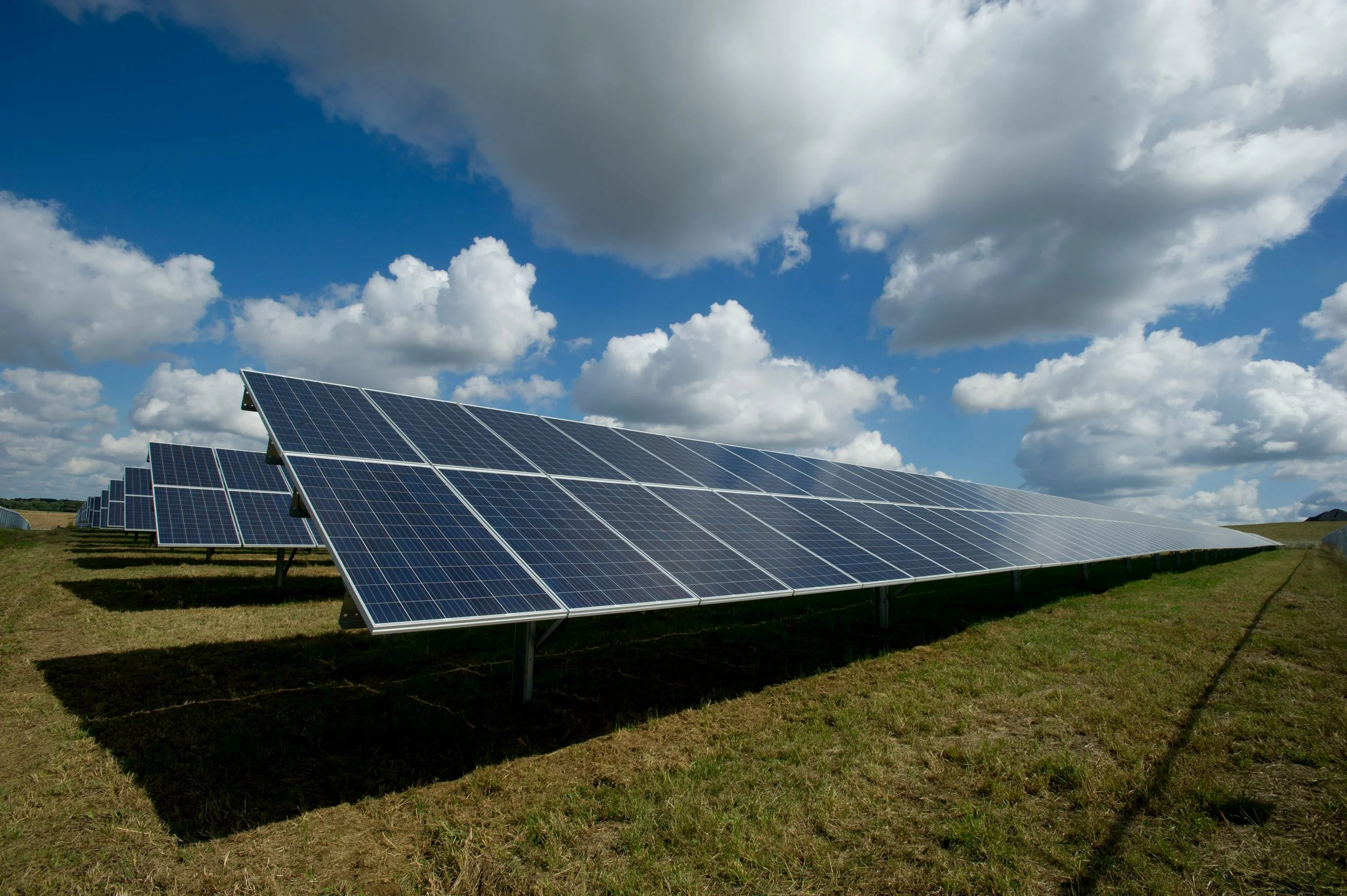 Multiple rows of solar panels installed on a grassy field under a partly cloudy sky.