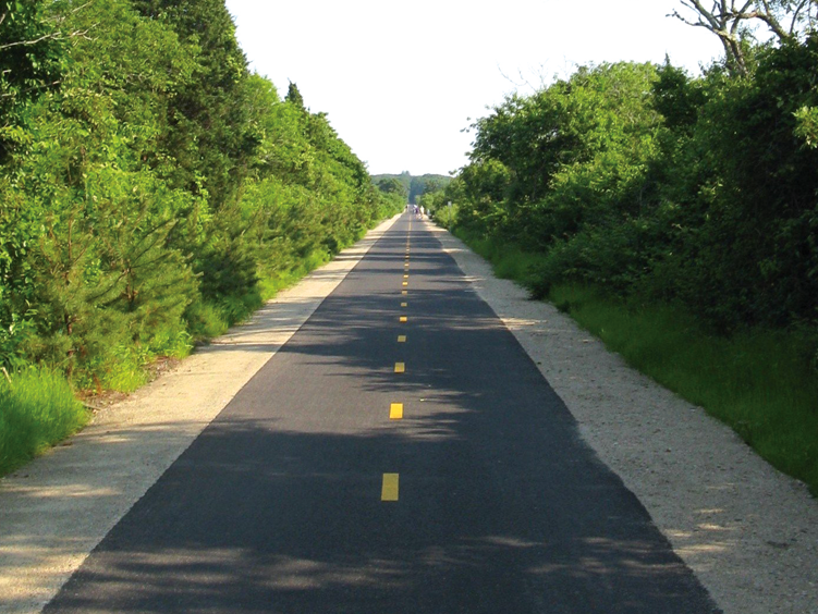 A straight narrow road with a yellow dashed centerline, surrounded by green trees and bushes on both sides.
