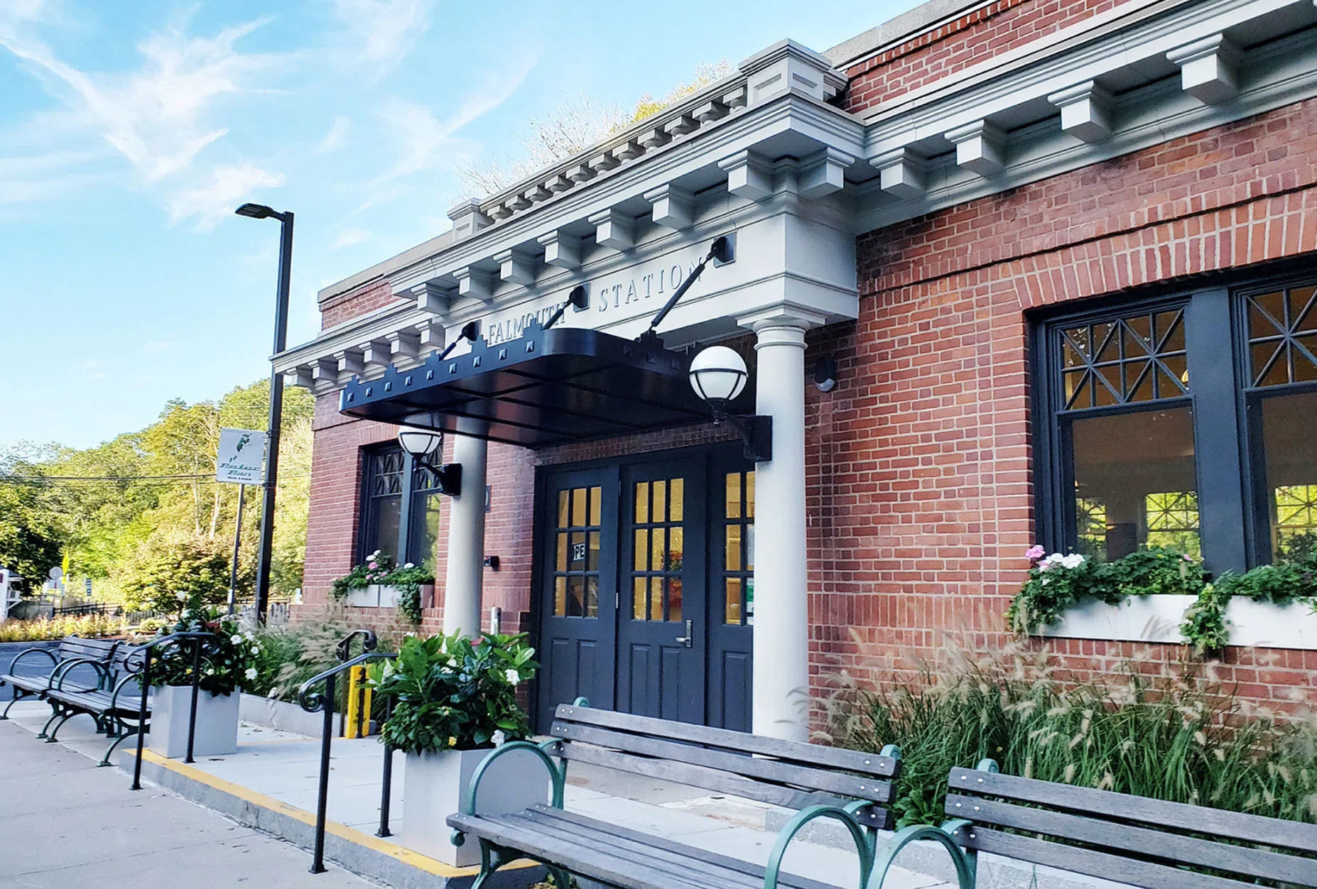 Exterior of a small town train station with brick walls, white columns, a black canopy, and black-framed windows; benches and flower planters are in the foreground.