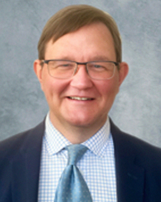 A smiling man with glasses and light brown hair, wearing a dark suit, blue checkered shirt, and a blue tie, standing against a gray background.