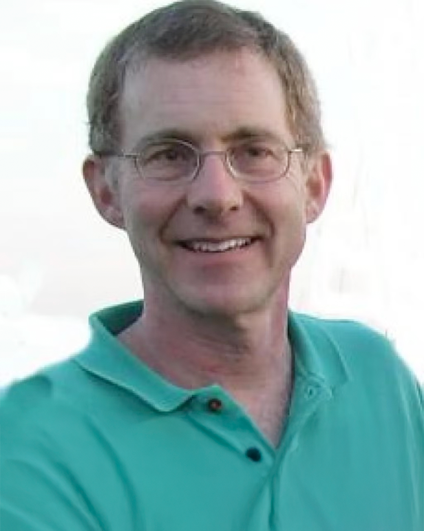 A man with glasses and short brown hair, smiling, wearing a turquoise polo shirt, outdoors with a bright background.