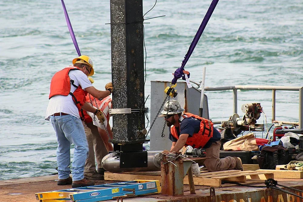 Three workers in safety vests and helmets are repairing or installing equipment on a pole in the water, on a platform over the water.