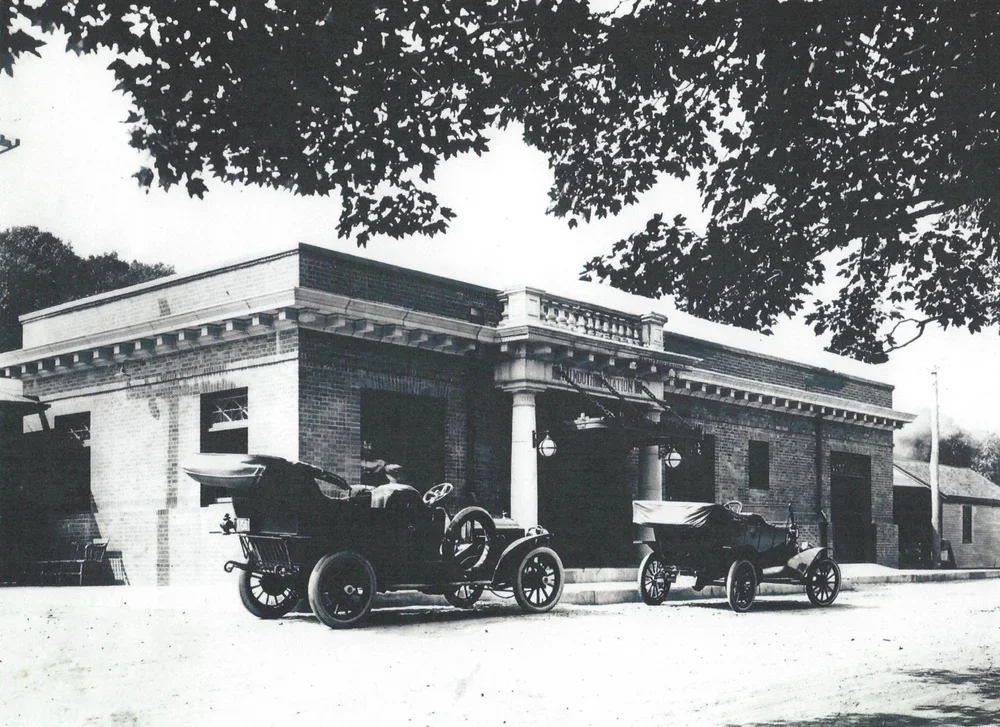 Black and white photo of a historic brick building labeled 'Chamber of Commerce,' with two vintage cars parked in front and trees overhead.