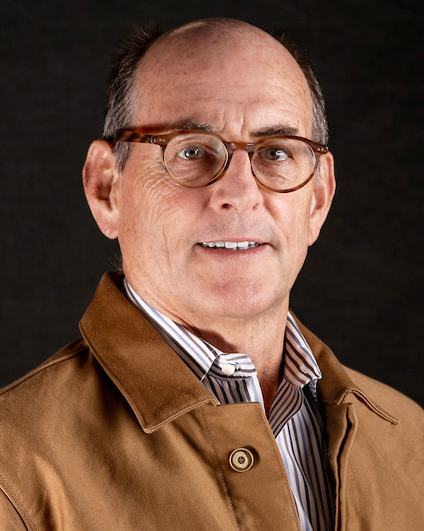 Portrait of a middle-aged man with glasses, wearing a striped shirt and a brown jacket against a dark background.