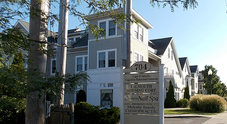 A street scene featuring a multi-story residential building with a sign in the foreground listing local businesses, including Cape & Islands Cognitive Behavioral Institute, Falmouth Housing Corp., Cranberry Nail Spa, and Mulcahy Family Chiropractic.