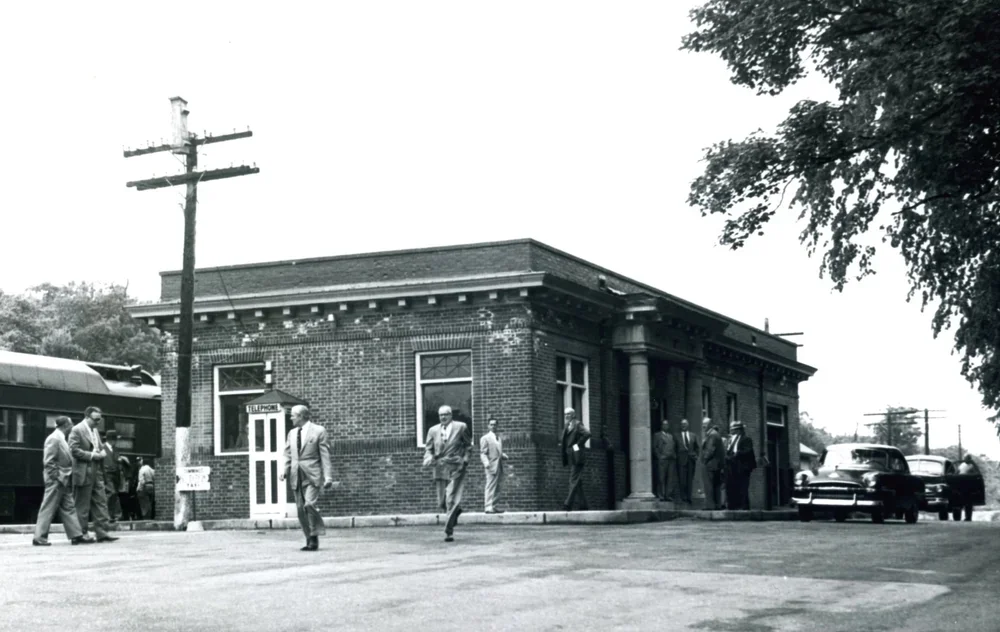 A black-and-white photograph of a small brick building with a few men in suits standing outside. There's a telephone booth next to the building and an electric pole nearby. A few cars and a bus are parked on the street.