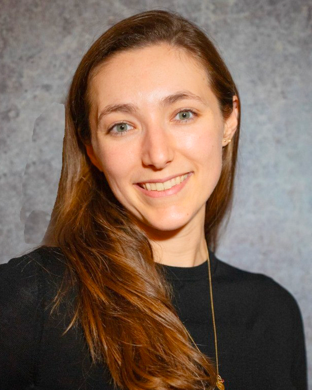A young woman with long, wavy brown hair smiling at the camera, wearing a black top and a necklace, against a gray textured background.
