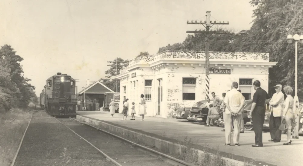 Black and white photo of a small train station with people waiting on the platform, a train arriving on the left, and a building with a sign that says 'Falmouth'.
