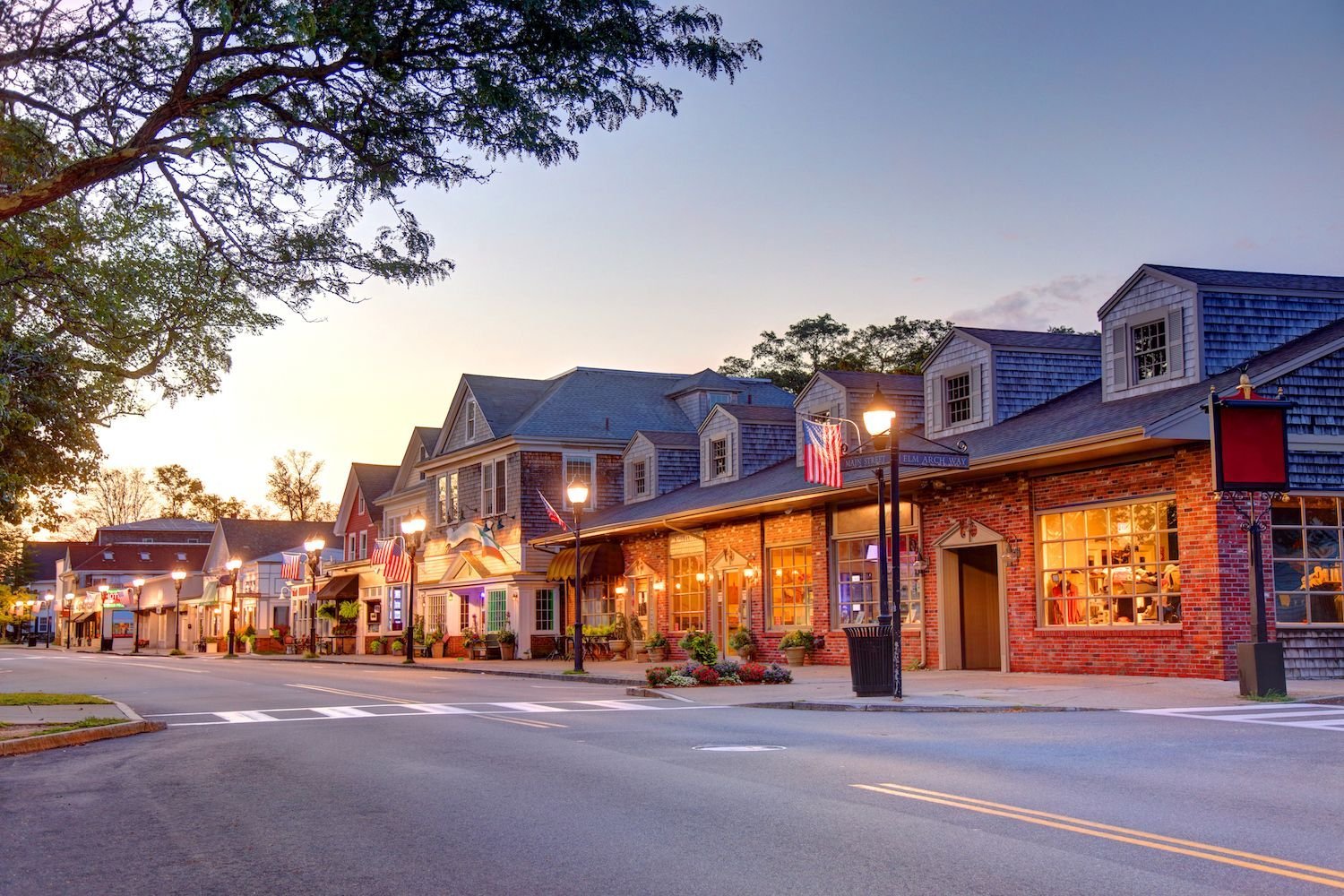 A small town street at sunset with cozy storefronts, American flags, and street lamps