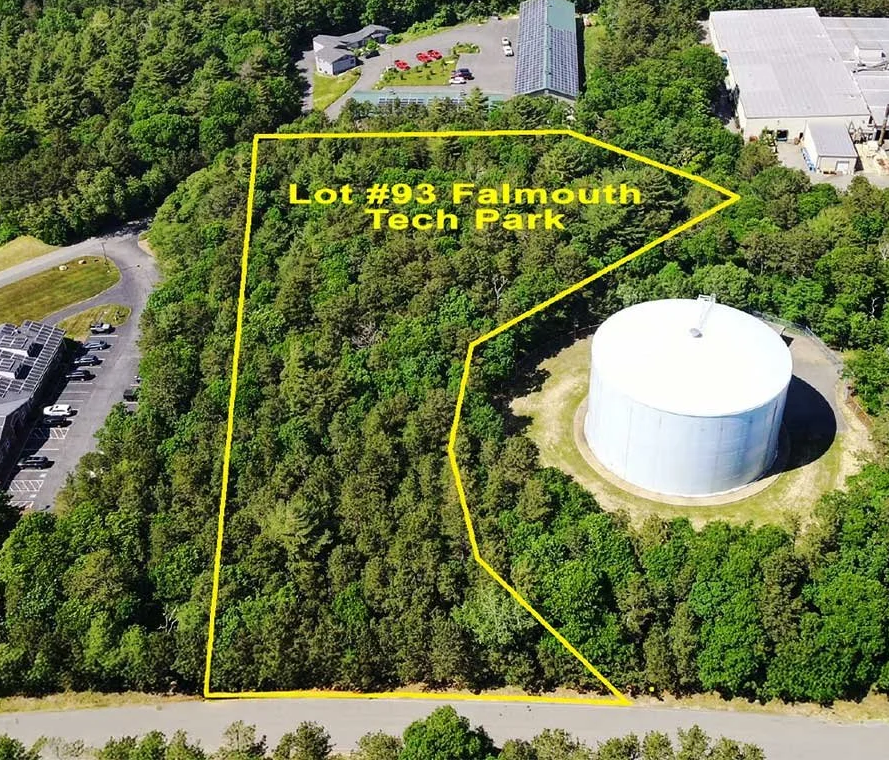 Aerial view of a wooded lot labeled 'Lot #93 Falmouth Tech Park' outlined in yellow, adjacent to a round white storage tank, parking lot with solar panels, and buildings surrounded by trees.