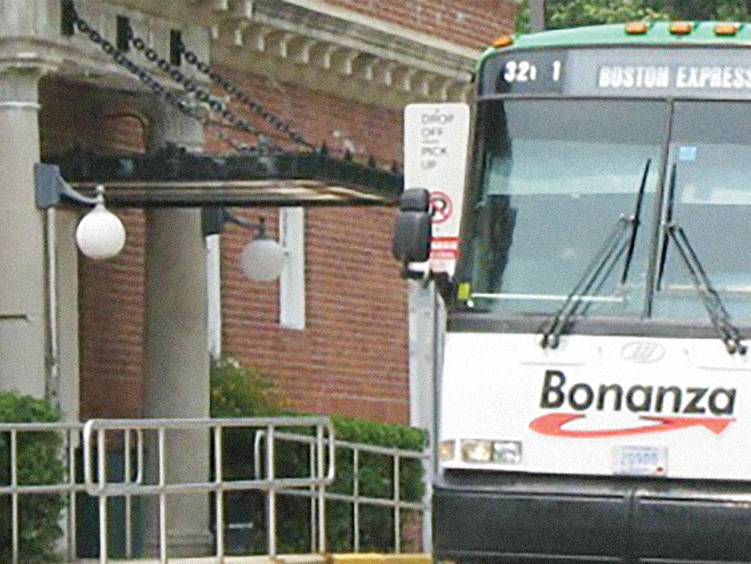 Front of a Boston Express bus with the Bonanza logo parked at a bus stop. The bus is near a building with a brick wall and decorative lighting.