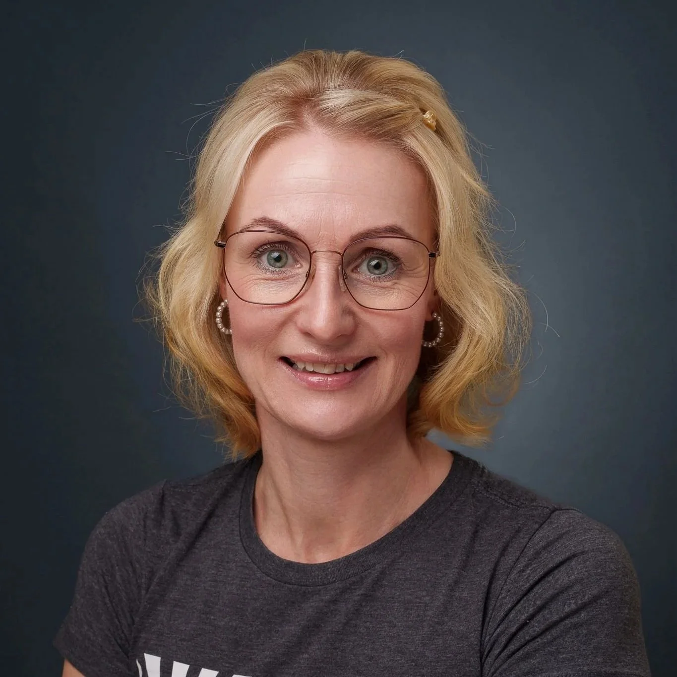 Porträt einer lachenden Frau mit blonde, lockiger Kurzhaarfrisur, Brille, Ohrringen und grauem T-Shirt vor dunklem Hintergrund.