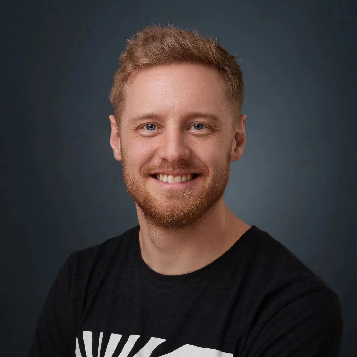 Ein junger Mann mit rotem Haar, Bart und einem schwarzen T-Shirt, lächelt vor dunklem Hintergrund.