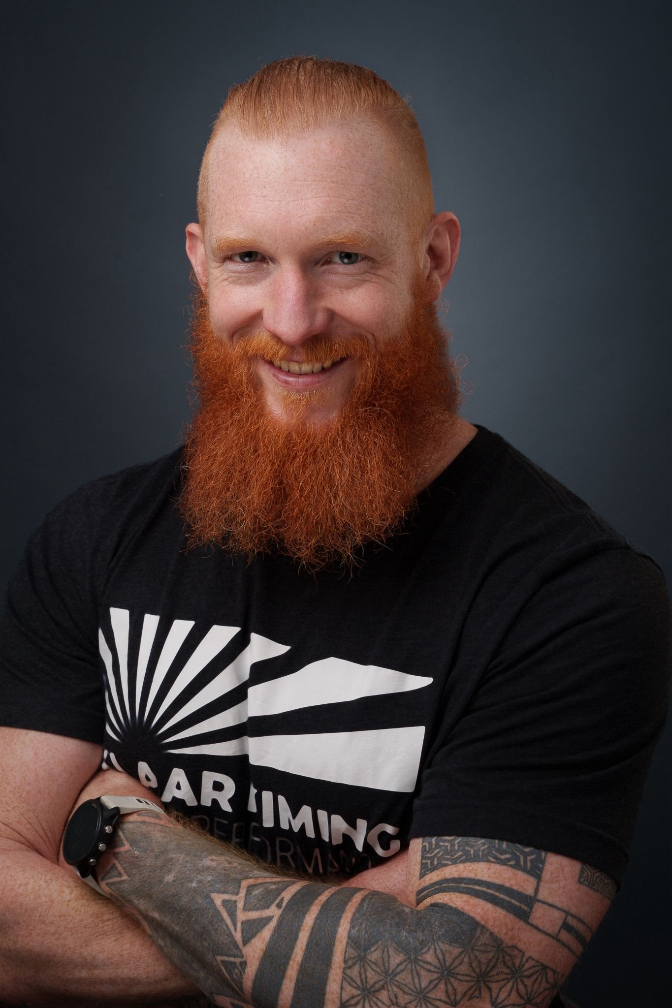 Ein Mann mit rotem Haart und Bart, trägt ein schwarzes T-Shirt mit weißen Linien und Text. Er lächelt, hat Tattoos am Arm, trägt einen schwarzen Armband und ist vor einem dunklen Hintergrund.