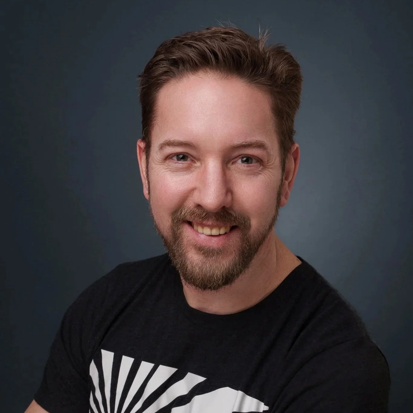 Porträt eines lächelnden jungen Mannes mit braunem Haar, Bart, dunklem T-Shirt, vor dunklem Hintergrund.