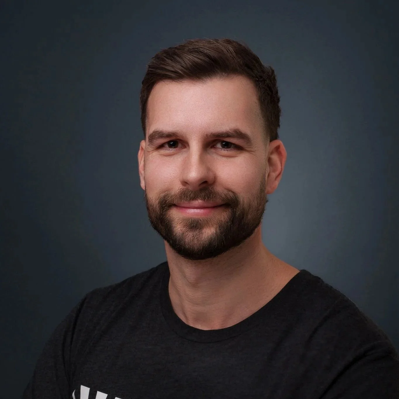Porträt eines lachenden Mannes mit dunklem Haar und Bart vor dunkelblauem Hintergrund, trägt ein schwarzes T-Shirt.