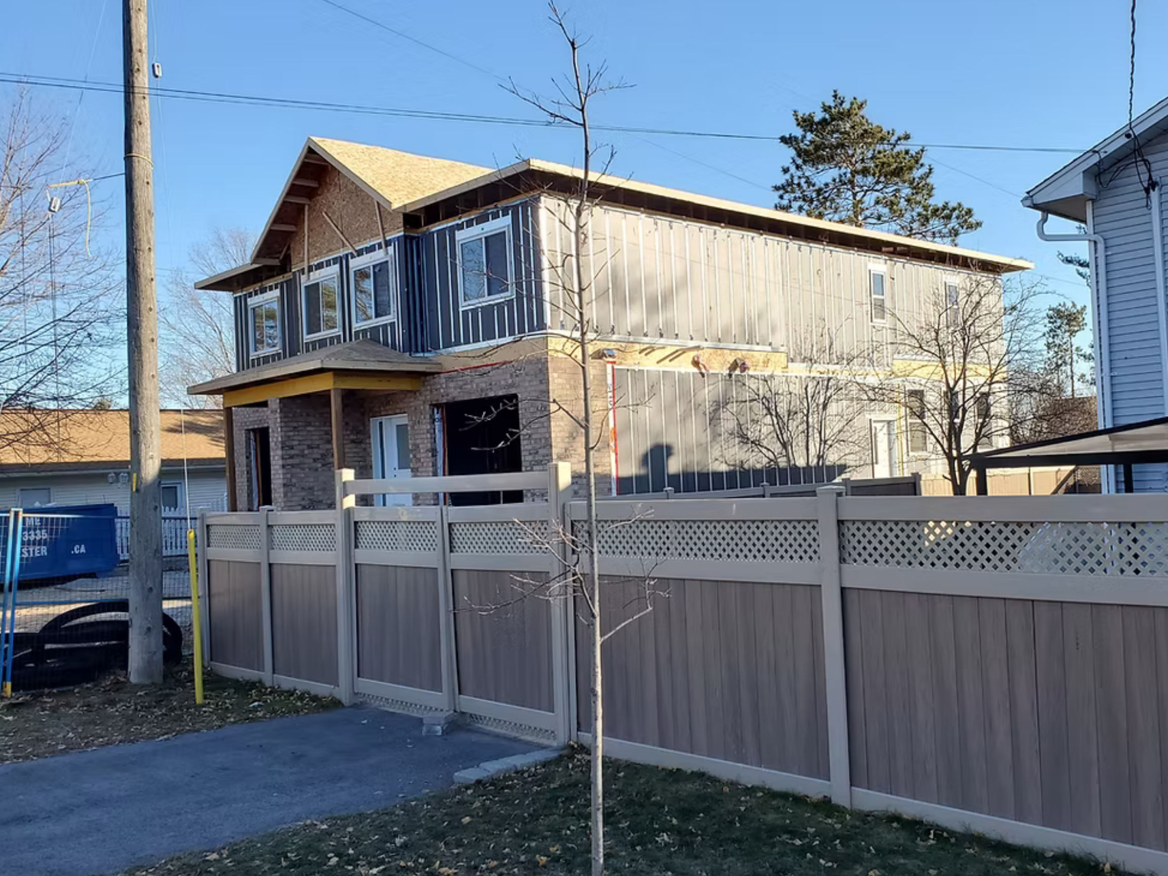 A side exterior view of a urban duplex build with the HARMONIC building envelope showing a large brown fence and sunny sky.