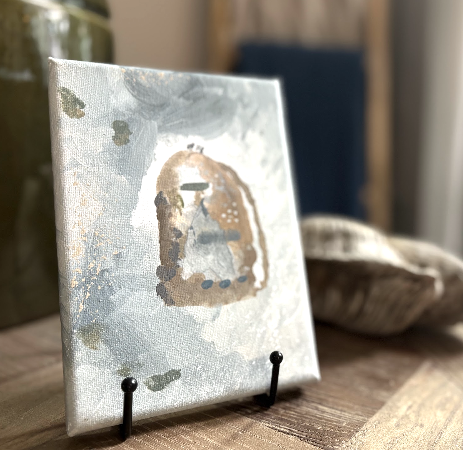 A small canvas with abstract, earthy-toned artwork held upright on a black stand. The background shows a blurred interior with wooden and fabric elements.