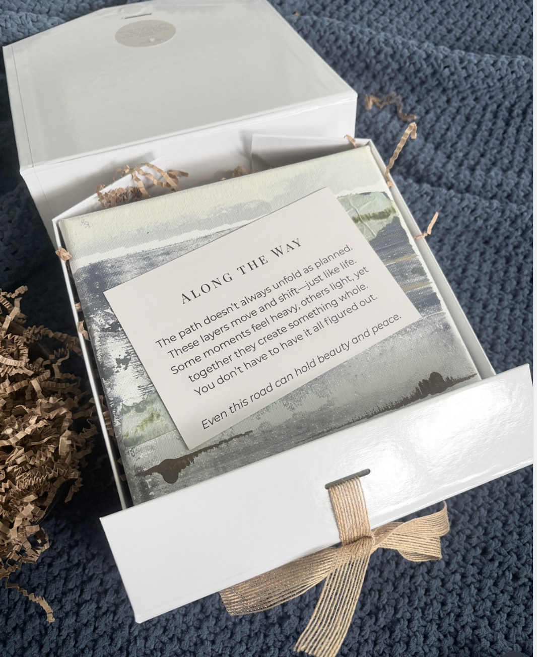 Open white box with shredded paper and a card inside, containing poetic text about a journey or path. The box is on a dark blue textured surface.