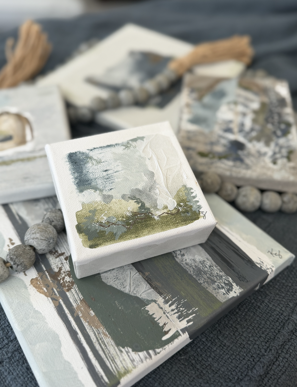 Small canvas painting of abstract trees and a landscape, set among other abstract landscape artworks and decorative objects on a textured surface in neutral blue, greens, beige, grays, whites and ivory..
