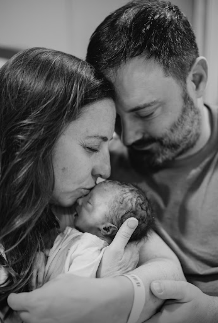 Black and white photo of a new mother, father, and their newborn baby in a hospital. The mother is holding the baby close to her face, the father is smiling with his forehead touching the mother's, all showing tenderness and love.