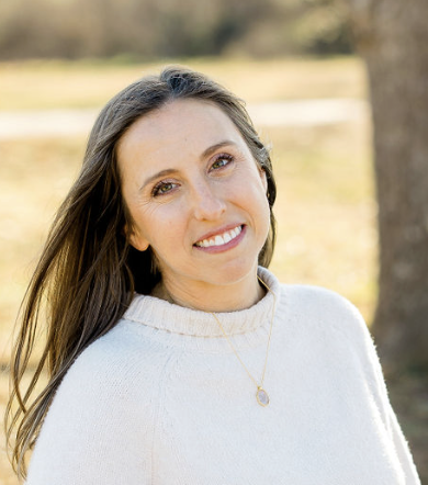 Smiling woman with long brown hair, wearing a cream sweater and a gold necklace, outdoors in a park.