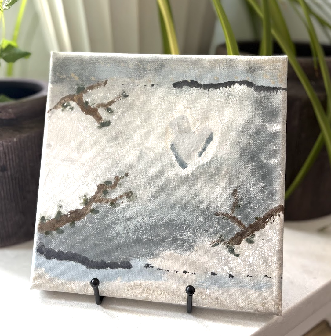 A small canvas painting depicts a heart shaped cloud in the sky with tree branches below, set against a soft sky background.