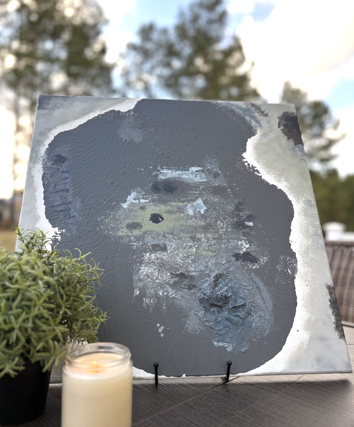 A square painting of a person's face with dark hair, using abstract textures and shades of gray, white, and black, displayed on a stand outdoors. There is a small potted plant and a lit candle in front of the painting, with trees and sky in the background.