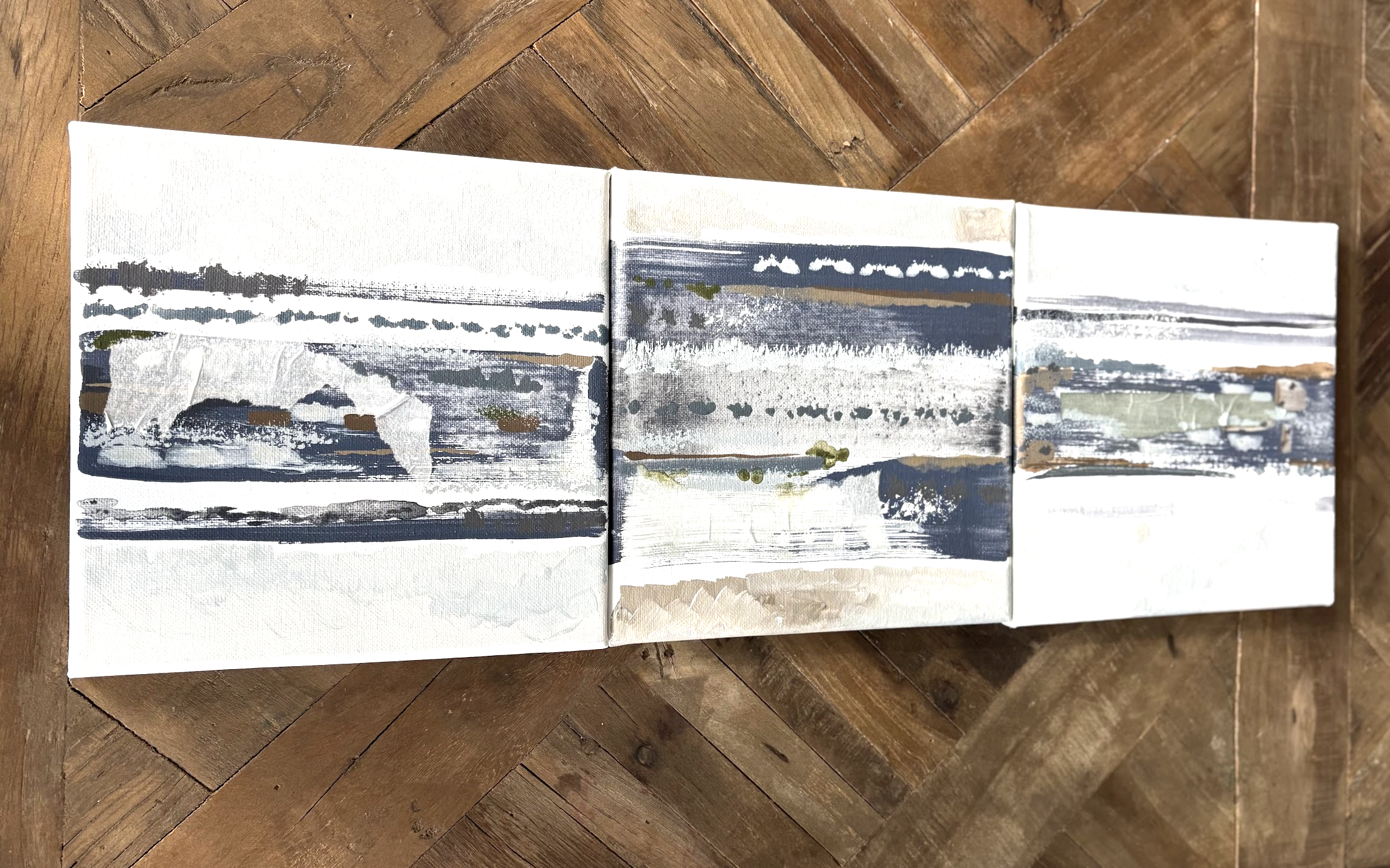 Three abstract landscape paintings with horizontal brush strokes in muted colors, placed side by side on a wooden surface.