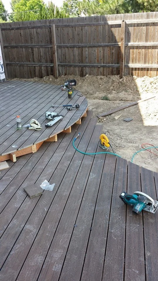 View of a backyard deck construction site with ongoing woodworking, power drills, and tools on the wooden deck, surrounded by a wooden fence and dirt ground.