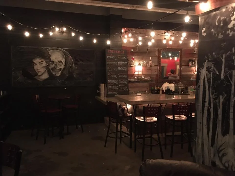Dimly lit bar or restaurant with string lights, a bartender, wooden walls, artwork of a woman and a skull, and a chalkboard menu with cocktails.