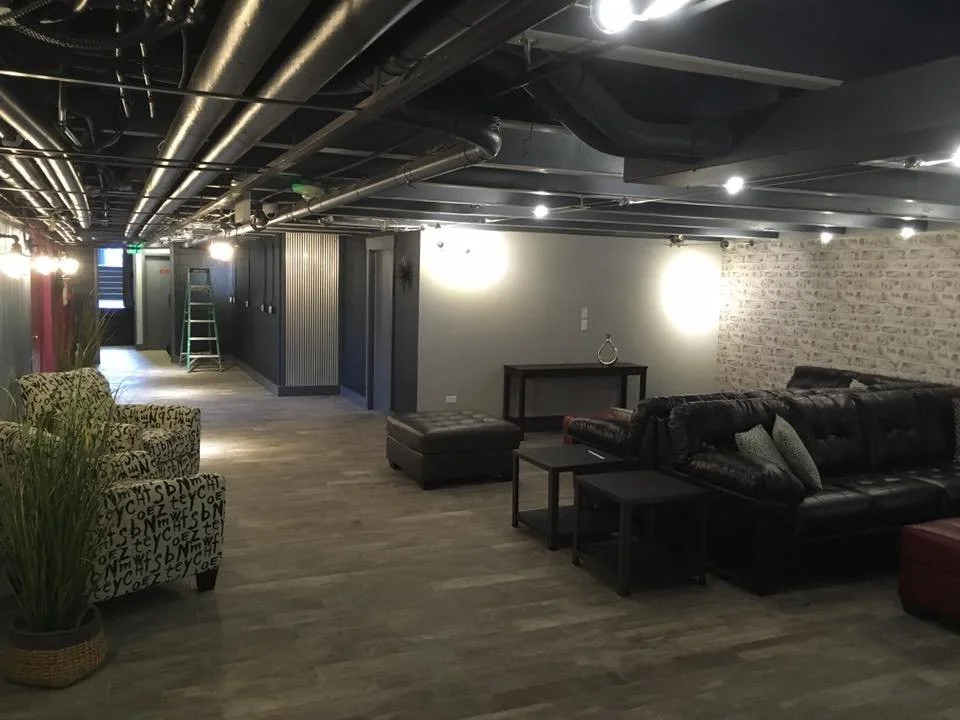 Basement lounge area with modern furniture, exposed ductwork, and brick wall.