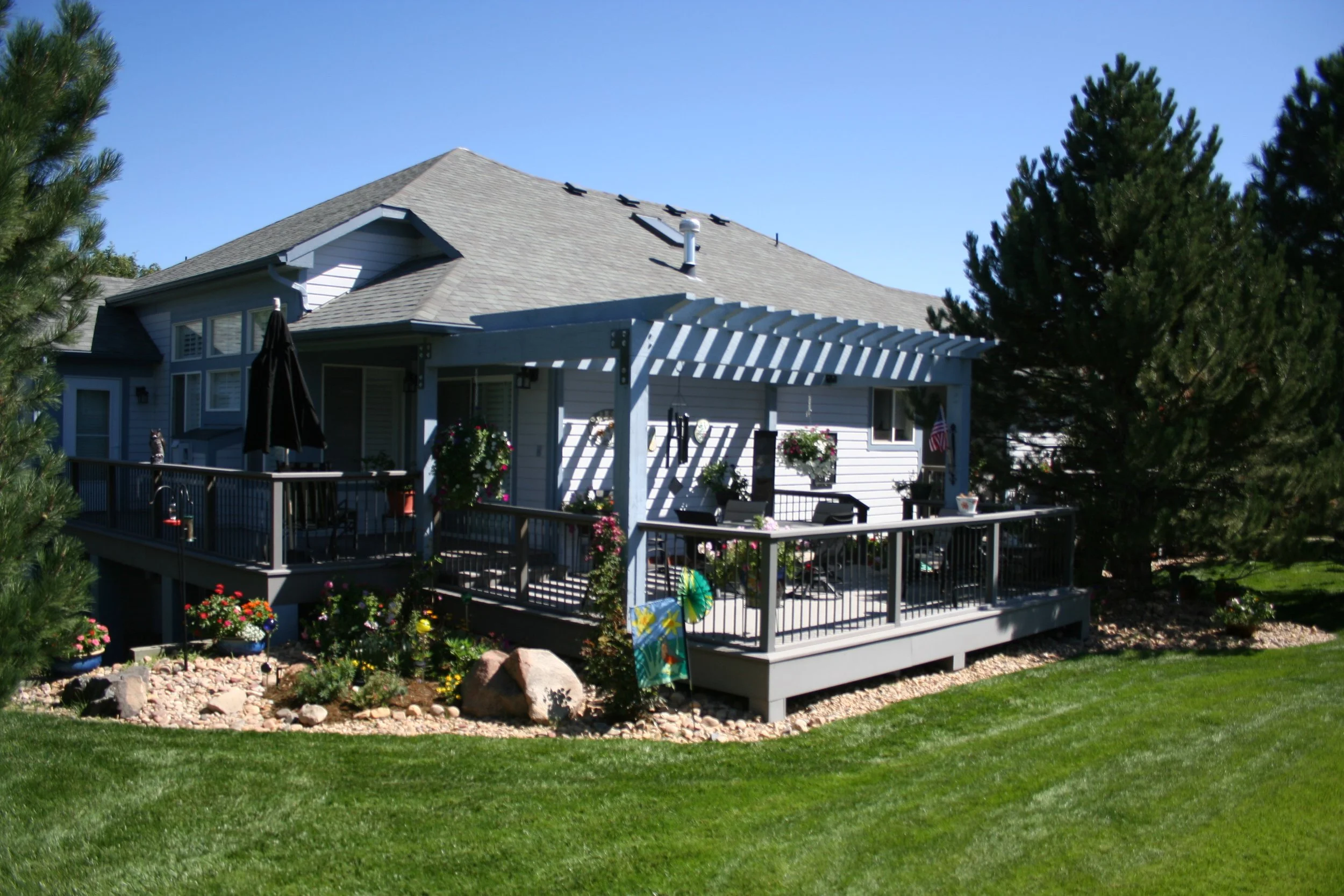 Backyard of a house featuring a wooden deck with furniture and a pergola, surrounded by well-maintained grass and landscaped garden with rocks and flowers, and tall trees in the background.